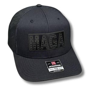 May include: Black trucker hat with a black patch that says "MAGA" with a stars and stripes pattern. The hat is made by Richardson and is adjustable.