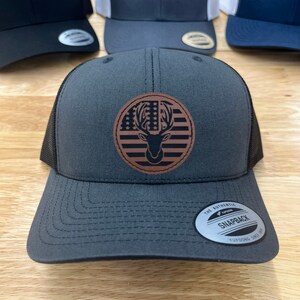 May include: A dark gray and black trucker hat with a brown leather patch featuring a deer silhouette and an American flag design. The patch has the text "Yupoong" on it. The hat has the text "The Authentic Yupoong Snapback Yupoong Since 1974" on the back.