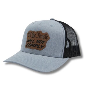 May include: A gray and black trucker hat with a brown leather patch that says "We the People Will Not Comply".