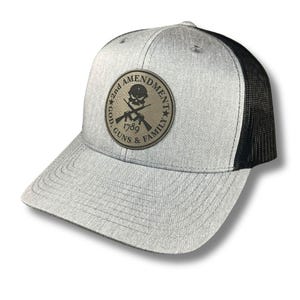 May include: A gray and black trucker hat with a brown leather patch that says "2nd Amendment God Guns & Family 1789" with a skull and crossbones design.