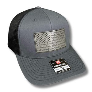 May include: A gray and black trucker hat with a silver metal patch featuring the Pledge of Allegiance and a stylized American flag. The hat is adjustable and has the Richardson logo on the back.