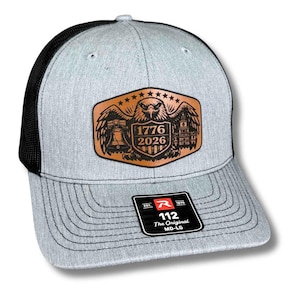May include: Gray trucker hat with a black mesh back and a curved brim. The front features a brown leather patch with an eagle, the Liberty Bell, and the text "1776 2026". A black tag with white text is on the brim.
