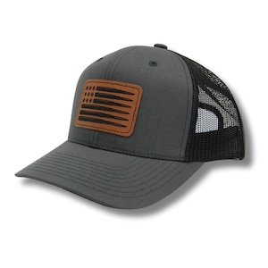May include: A gray and black trucker hat with a brown leather patch featuring a US flag design made of bullet casings.