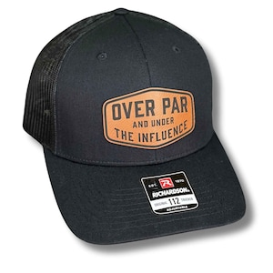 May include: Black trucker hat with a mesh back and a curved brim. The hat features a brown leather patch with the words "OVER PAR AND UNDER THE INFLUENCE". The hat has an adjustable strap and a Richardson 112 tag.