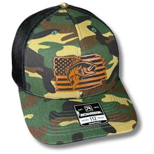 May include: A camouflage trucker hat with a black mesh back and a brown leather patch on the front. The patch features an American flag design with a fish graphic. A black and white sticker on the brim reads: EST. 1970 RICHARDSON ORIGINAL 112 TRUCKER ADJUSTABLE.
