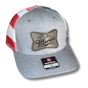 May include: Gray trucker hat with a silver patch that reads "Merica 1776" with an eagle and mountain design. The hat features a red, white, and blue American flag design on the side and a Richardson 112 Trucker tag.