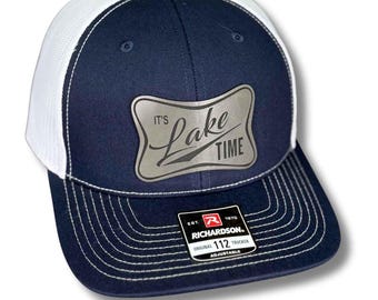 May include: Navy blue trucker hat with a white mesh back and a curved brim. The hat features a silver patch that reads "It's Lake Time." The Richardson 112 hat is adjustable.