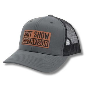 May include: A gray and black trucker hat with a brown leather patch that says "Shit Show Supervisor".