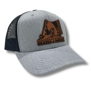 May include: A gray and black trucker hat with a brown leather patch featuring a group of people raising a flag with the text "Grazed & Unfazed".