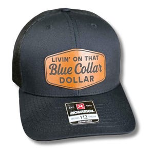 May include: Black trucker hat with a mesh back and a curved brim. The hat features a brown leather patch with the words "LIVIN' ON THAT Blue Collar DOLLAR". The hat also has a Richardson tag.