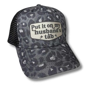 May include: A black and white leopard print trucker hat with a black mesh back. The hat has a silver patch with the text "Put it on my husband's tab" in black.