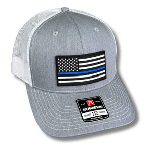 May include: Gray trucker hat with a white mesh back and a curved brim. The front features a patch of the American flag with a blue stripe. The hat includes a Richardson tag and is adjustable.