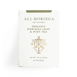 May include: A white box with gold lettering that reads "ALL MORINGA 100% NATURAL ORGANIC MORINGA LEAF & MINT TEA". The box also features a gold logo of a stylized tree and the text "Net WT 1.76 oz (50 g) 28 TEA BAGS".
