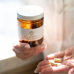 May include: A brown glass bottle with a white label that reads "ALL MORINGA PURE MORINGA LEAF CAPSULES". Two green capsules are in the palm of a hand.
