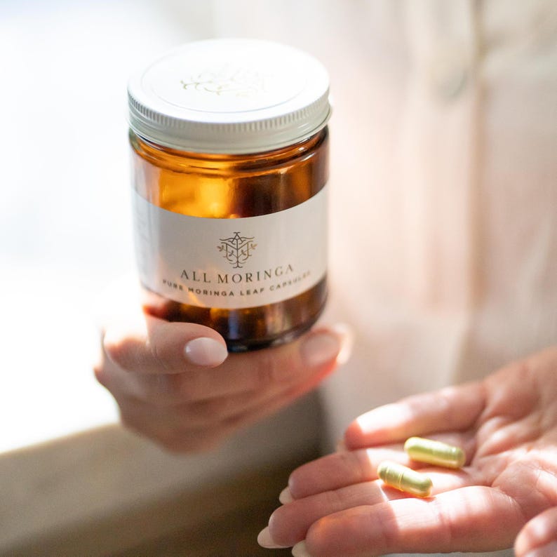 May include: A brown glass bottle with a white label that reads "ALL MORINGA PURE MORINGA LEAF CAPSULES". Two green capsules are in the palm of a hand.