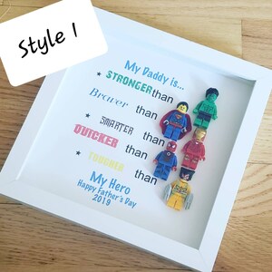 May include: A white frame with a blue and white background. The text "My Daddy is..." is written in blue. The text "STRONGER than" is written in red. The text "Braver than" is written in blue. The text "SMARTER than" is written in red. The text "QUICKER than" is written in red. The text "TOUGHER than" is written in red. The text "My Hero" is written in yellow. The text "Happy Father's Day 2019" is written in yellow. There are six miniature superhero figurines in the frame.