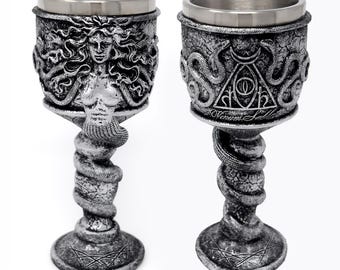 May include: Two silver-toned goblets with metallic finishes. One features a relief of a figure with snake hair, while the other displays a triangular symbol with an eye and the text Venum Sabla. Both have snake-wrapped stems and circular bases.