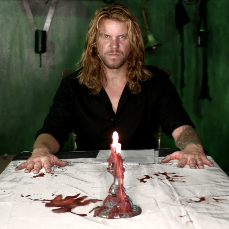 Puede incluir: Un hombre con el pelo largo y rubio se sienta a una mesa con un mantel blanco. La mesa est&aacute; cubierta de salpicaduras de sangre. Una vela roja est&aacute; encendida en un candelabro de plata.