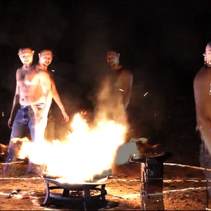 Puede incluir: Cuatro hombres sin camisa se paran alrededor de una fogata en un entorno oscuro. Los hombres llevan pantalones vaqueros y miran el fuego. El fuego es grande y brillante, con llamas que alcanzan lo alto en el aire.