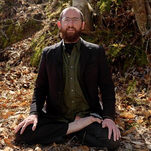 Puede incluir: Una persona con gafas, una camisa verde abotonada y una blazer oscura est&aacute; sentada con las piernas cruzadas al aire libre. La persona tiene barba y est&aacute; rodeada de hojas ca&iacute;das y follaje.