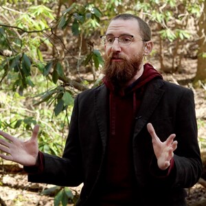 Puede incluir: Una persona con gafas, una sudadera con capucha burdeos y una blazer gris oscuro. La persona tiene una larga barba marr&oacute;n y est&aacute; de pie en una zona boscosa con follaje verde en el fondo. La persona gesticula con ambas manos.