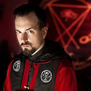 Puede incluir: Un hombre con barba de chivo lleva una t&uacute;nica roja y negra con un s&iacute;mbolo de pentagrama en el pecho. Est&aacute; de pie frente a un fondo rojo con un s&iacute;mbolo de pentagrama.