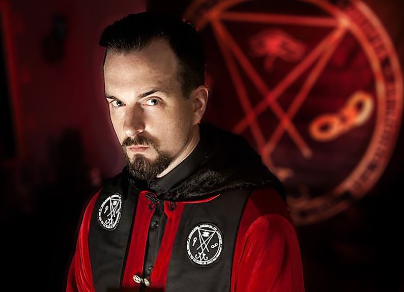 Puede incluir: Un hombre con barba de chivo lleva una t&uacute;nica roja y negra con un s&iacute;mbolo de pentagrama en el pecho. Est&aacute; de pie frente a un fondo rojo con un s&iacute;mbolo de pentagrama.