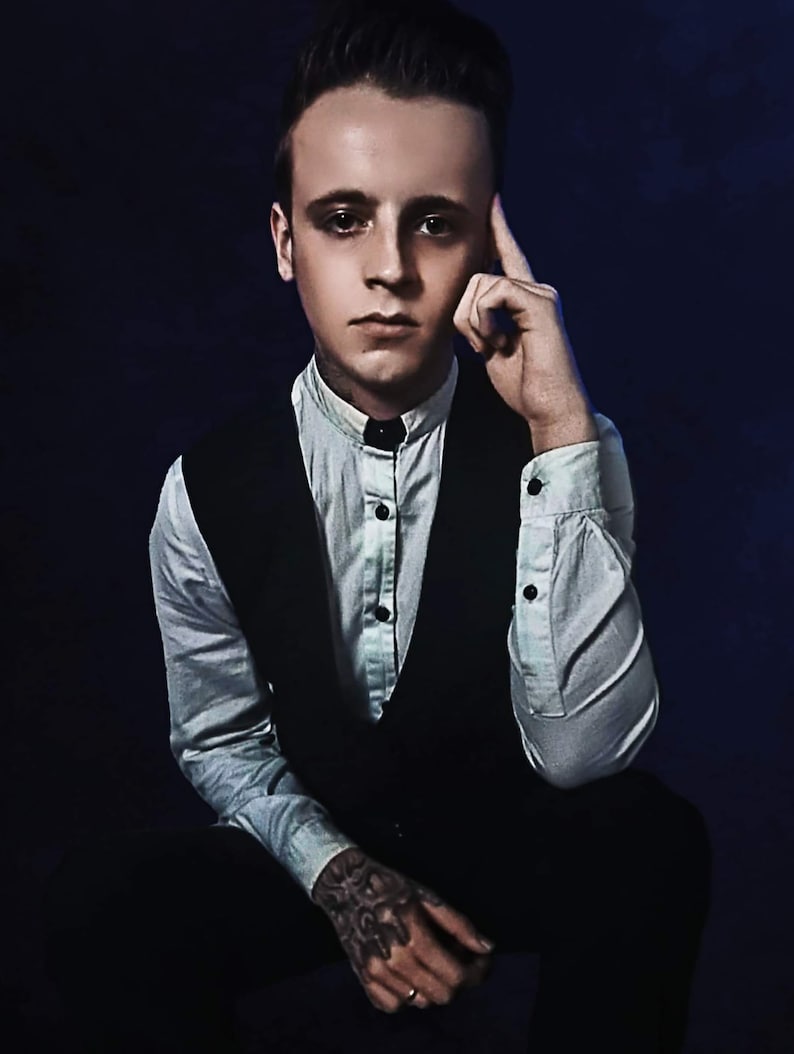 Puede incluir: Un hombre con una camisa blanca abotonada y un chaleco negro. Est&aacute; sentado con la mano en la barbilla y mirando a la c&aacute;mara.