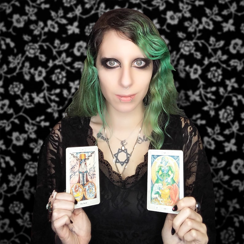 Puede incluir: Dos cartas de tarot, El Diablo y La Emperatriz, son sostenidas por una persona con cabello verde. La carta del Diablo presenta una figura roja y negra con cuernos y cola. La carta de la Emperatriz presenta una mujer con un vestido verde con una corona y un orbe dorado.