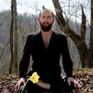 Puede incluir: Una persona con un traje negro est&aacute; sentada con las piernas cruzadas al aire libre, sosteniendo un narciso amarillo. La persona tiene barba y gafas. El fondo es un bosque con &aacute;rboles desnudos y hojas ca&iacute;das.