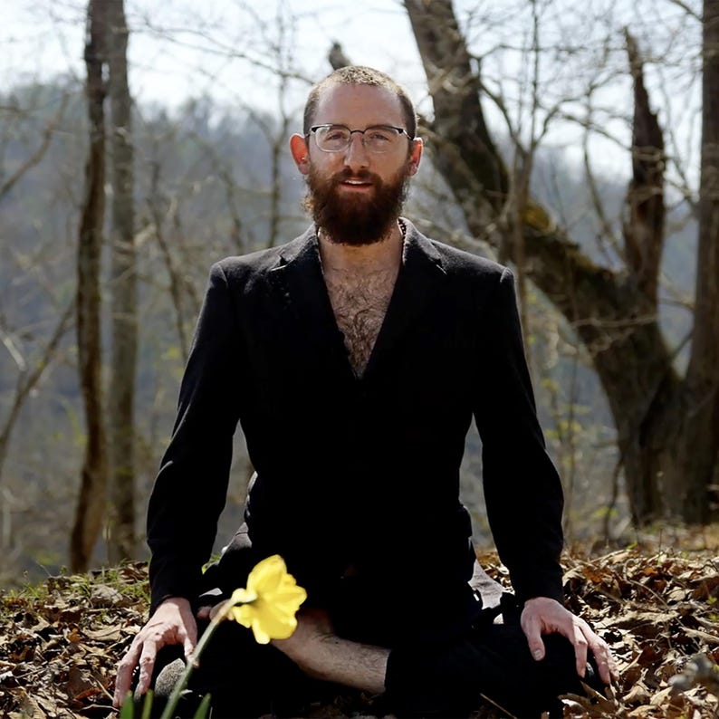 Puede incluir: Una persona con un traje negro est&aacute; sentada con las piernas cruzadas al aire libre, sosteniendo un narciso amarillo. La persona tiene barba y gafas. El fondo es un bosque con &aacute;rboles desnudos y hojas ca&iacute;das.