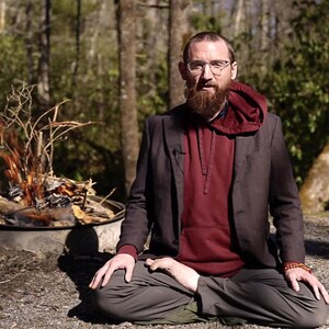 Puede incluir: Una persona con una sudadera con capucha granate y una blazer gris oscuro est&aacute; sentada con las piernas cruzadas al aire libre. La persona tiene una larga barba y gafas. Un peque&ntilde;o fuego arde en una hoguera de metal en el fondo, rodeado de &aacute;rboles.