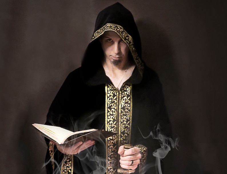 Puede incluir: Un hombre con una t&uacute;nica negra con capucha y ribetes dorados sostiene un libro abierto. La t&uacute;nica tiene intrincados bordados dorados y la capucha est&aacute; puesta sobre su cabeza. Sale humo del libro.