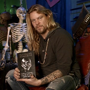 May include: A man with long blonde hair and a beard is sitting in a chair holding a black book with a silver skull design. The book's title is "Gateways to the Unseen World" by Edgar Cayce & The ARE. The man is wearing a black shirt and blue jeans. A skeleton is hanging in the background.