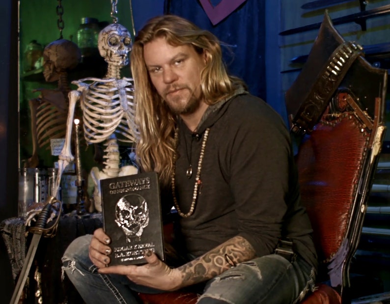 May include: A man with long blonde hair and a beard is sitting in a chair holding a black book with a silver skull design. The book's title is "Gateways to the Unseen World" by Edgar Cayce & The ARE. The man is wearing a black shirt and blue jeans. A skeleton is hanging in the background.