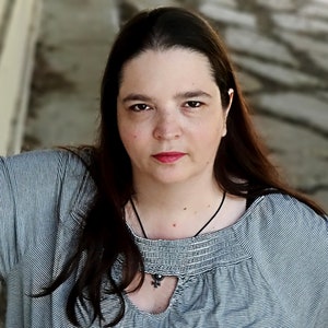 Puede incluir: Una mujer con el pelo largo y oscuro que lleva una camisa gris a rayas con un collar negro y un colgante.