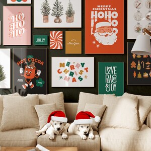 Op de afbeelding: Een woonkamer die is versierd voor kerstmis met een bank, twee witte puppy's met kerstmanmutsen en verschillende kerstthemaposters aan de muur. De posters bevatten zinnen als "Merry Christmas", "Ho Ho Ho", "Love Peace & Joy" en "Hot Cocoa".