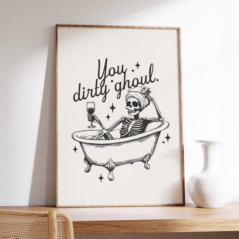 Funny Halloween decor. Light beige background with black foreground. Illustration of a skeleton sitting in a vintage bathtub with a glass of wine and text that reads you dirty ghoul. Perfect bathroom decor for the fall autumn season.