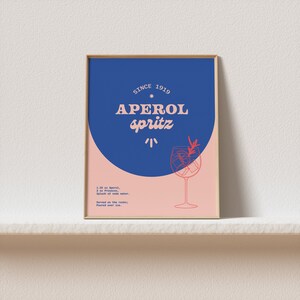 May include: A framed poster with a blue and pink background. The poster features a stylized illustration of a glass of Aperol Spritz with the text "SINCE 1919 APEROL spritz" in red and white. The poster also includes the recipe for the cocktail: 1.25 oz Aperol, 2 oz Prosecco, Splash of soda water. Served on the rocks: Poured over ice.