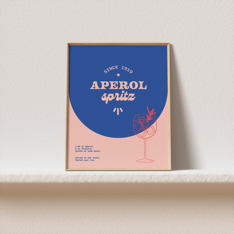 May include: A framed poster with a blue and pink background. The poster features a stylized illustration of a glass of Aperol Spritz with the text "SINCE 1919 APEROL spritz" in red and white. The poster also includes the recipe for the cocktail: 1.25 oz Aperol, 2 oz Prosecco, Splash of soda water. Served on the rocks: Poured over ice.