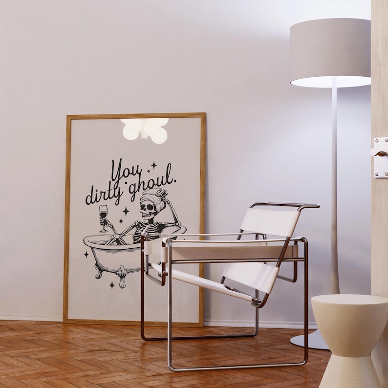 Funny Halloween decor. Light beige background with black foreground. Illustration of a skeleton sitting in a vintage bathtub with a glass of wine and text that reads you dirty ghoul. Perfect bathroom decor for the fall autumn season.