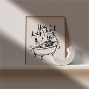Funny Halloween decor. Light beige background with black foreground. Illustration of a skeleton sitting in a vintage bathtub with a glass of wine and text that reads you dirty ghoul. Perfect bathroom decor for the fall autumn season.