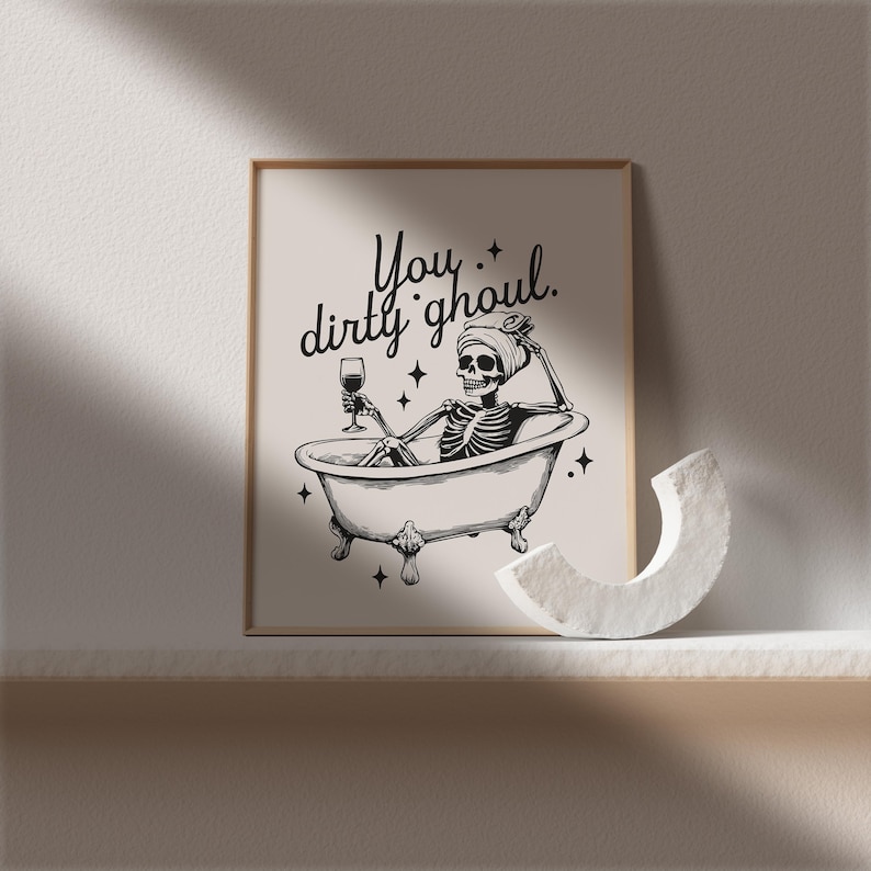 Funny Halloween decor. Light beige background with black foreground. Illustration of a skeleton sitting in a vintage bathtub with a glass of wine and text that reads you dirty ghoul. Perfect bathroom decor for the fall autumn season.