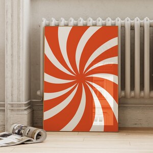 Op de afbeelding: Een ingelijst afdruk met een rood en wit spiraalontwerp. De afdruk hangt aan een witte muur naast een witte radiator.
