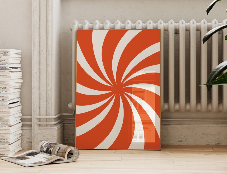 Op de afbeelding: Een ingelijst afdruk met een rood en wit spiraalontwerp. De afdruk hangt aan een witte muur naast een witte radiator.