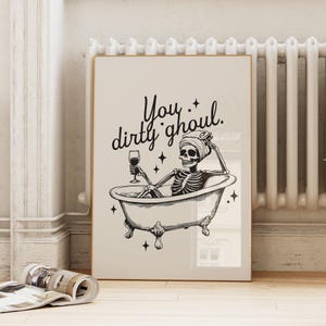 Funny Halloween decor. Light beige background with black foreground. Illustration of a skeleton sitting in a vintage bathtub with a glass of wine and text that reads you dirty ghoul. Perfect bathroom decor for the fall autumn season.