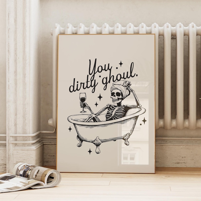 Funny Halloween decor. Light beige background with black foreground. Illustration of a skeleton sitting in a vintage bathtub with a glass of wine and text that reads you dirty ghoul. Perfect bathroom decor for the fall autumn season.