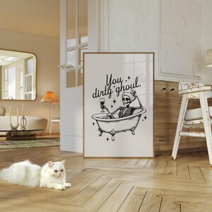 Funny Halloween decor. Light beige background with black foreground. Illustration of a skeleton sitting in a vintage bathtub with a glass of wine and text that reads you dirty ghoul. Perfect bathroom decor for the fall autumn season.