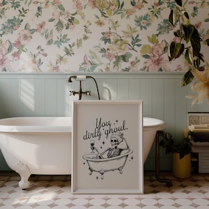 Funny Halloween decor. Light beige background with black foreground. Illustration of a skeleton sitting in a vintage bathtub with a glass of wine and text that reads you dirty ghoul. Perfect bathroom decor for the fall autumn season.