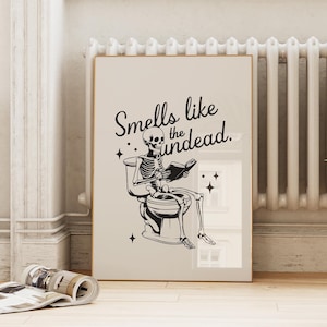 May include: A black and white illustration of a skeleton sitting on a toilet reading a book. The text "Smells like the undead." is above the skeleton.